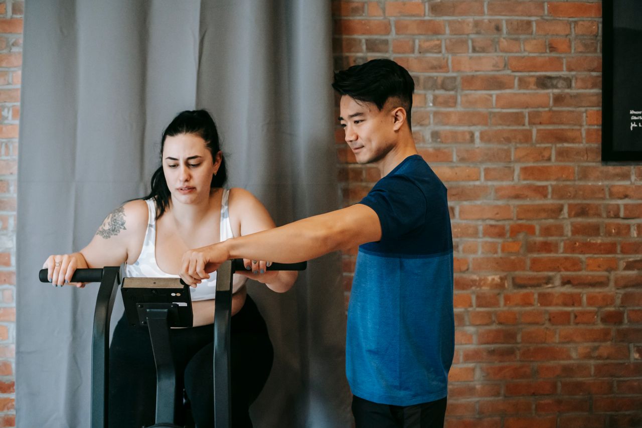 Ethnic personal fitness coach standing near overweight female training on exercise bike during cardio workout